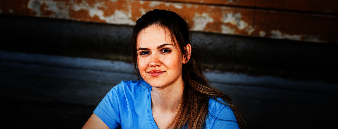 Eine junge Frau mit braunen langen Haaren und Pferdeschwanz sitzt auf einer Stiege, sie trägt ein hellblaues T-Shirt mit Caritas Logo und lächelt in die Kamera.