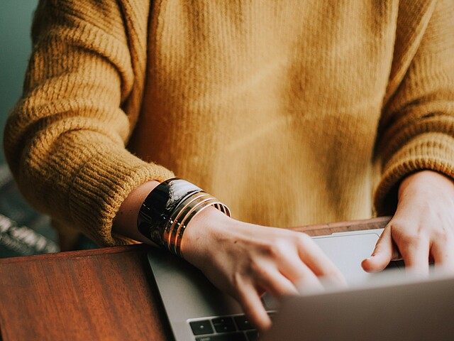 Eine junge Frau arbeitet am Laptop, ihre Hände auf der Tastatur sind im Fokus, ihr Gesicht nicht zu sehen.